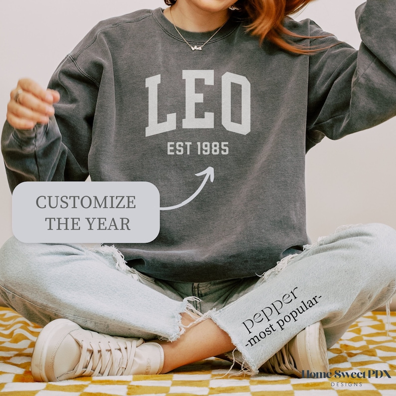 Puede incluir: Una sudadera gris con la palabra "LEO" en letras blancas y el texto "EST 1985" debajo. La sudadera la lleva puesta una persona sentada. El texto "CUSTOMIZE THE YEAR" es visible en un cuadro blanco.