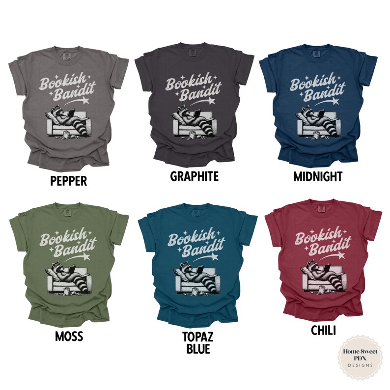 May include: Six different color t-shirts with the text "Bookish Bandit" and a cartoon raccoon reading a book on a couch. The colors are pepper, graphite, midnight, moss, topaz blue, and chili.