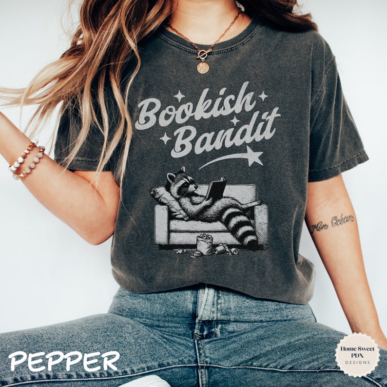May include: A dark gray t-shirt with the text "Bookish Bandit" in white lettering. A cartoon raccoon wearing a shirt is sitting on a couch reading a book. The raccoon is surrounded by snacks and a star.