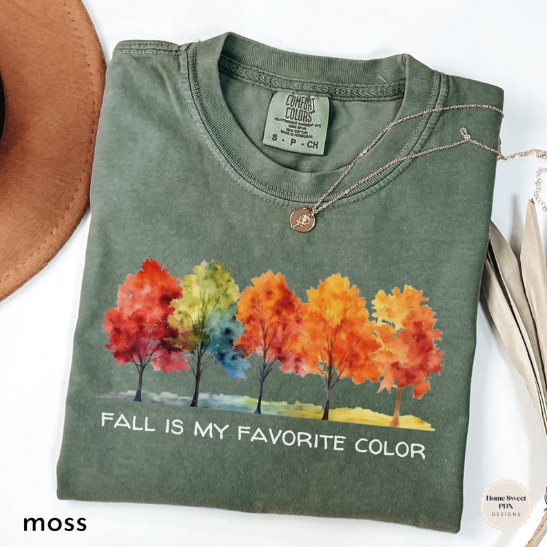 May include: A green t-shirt with a watercolor design of fall leaves in shades of red, orange, yellow, and green. The text "FALL IS MY FAVORITE COLOR" is printed in white on the shirt.