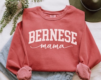 Puede incluir: Una sudadera roja con el texto "BERNESE mama" en letras blancas. La sudadera está doblada y se muestra sobre una superficie blanca con un par de pantalones vaqueros azules y un sombrero verde.
