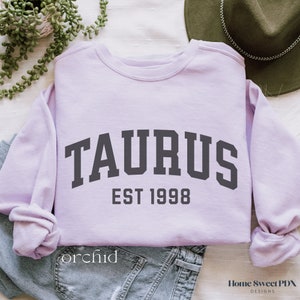 May include: A lavender sweatshirt with the text "TAURUS EST 1998" in dark gray.