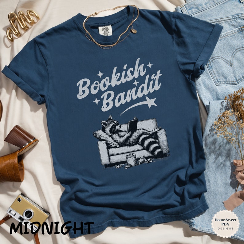 May include: A navy blue t-shirt with a white graphic of a raccoon reading a book on a couch. The text "Bookish Bandit" is written in a white, stylized font above the raccoon.
