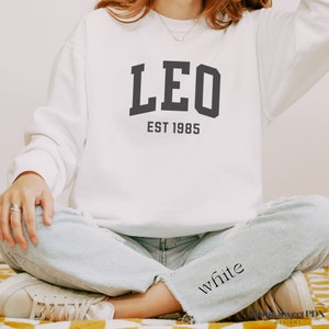 Puede incluir: Sudadera blanca con texto negro que dice "LEO EST 1985".