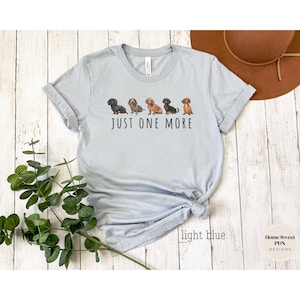 Puede incluir: Camiseta azul claro con una l&iacute;nea de cinco perros salchicha y el texto "JUST ONE MORE".