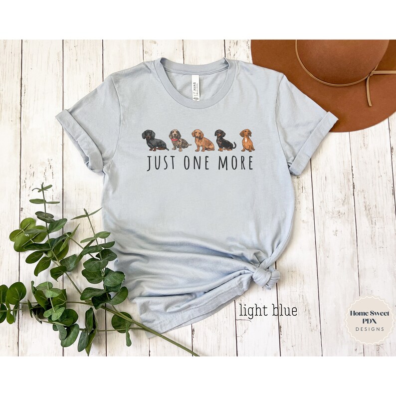 Puede incluir: Camiseta azul claro con una l&iacute;nea de cinco perros salchicha y el texto "JUST ONE MORE".