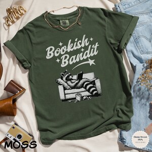 May include: A green t-shirt with a white graphic of a raccoon reading a book on a couch. The text "Bookish Bandit" is above the raccoon.