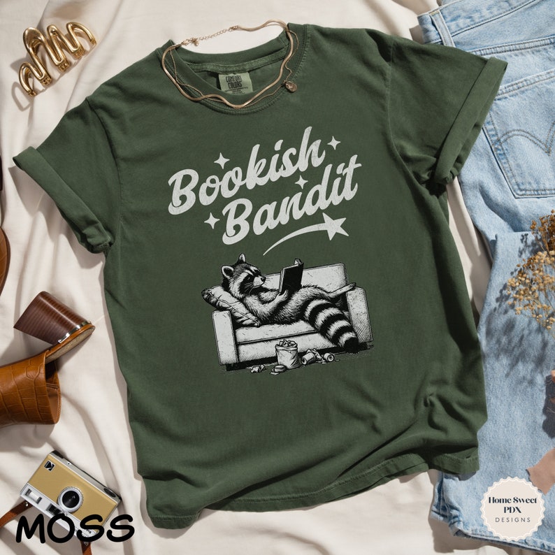 May include: A green t-shirt with a white graphic of a raccoon reading a book on a couch. The text "Bookish Bandit" is above the raccoon.
