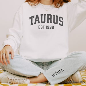 May include: White sweatshirt with the text "TAURUS EST 1998" in black lettering. The sweatshirt is being worn by a person sitting with their legs crossed.