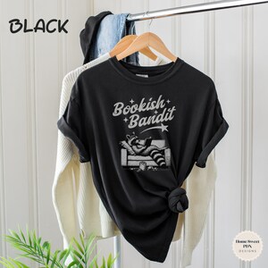May include: Black t-shirt with a graphic of a raccoon reading a book on a couch. The text "Bookish Bandit" is printed on the shirt.