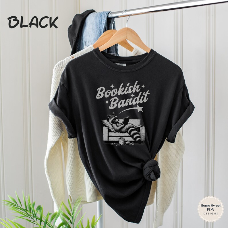 May include: Black t-shirt with a graphic of a raccoon reading a book on a couch. The text "Bookish Bandit" is printed on the shirt.