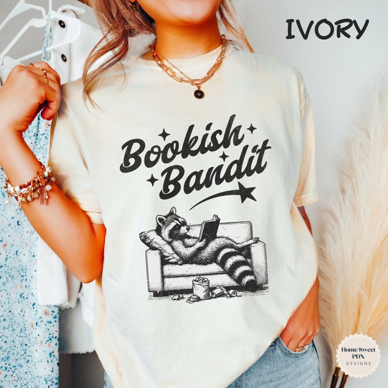 May include: A light ivory t-shirt with a black and white illustration of a raccoon reading a book on a couch. The text "Bookish Bandit" is printed in black on the shirt.