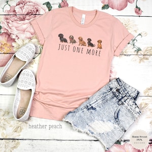 Puede incluir: Una camiseta de color melocot&oacute;n con una l&iacute;nea de cinco perros salchicha de diferentes colores y patrones. El texto "Just One More" est&aacute; impreso debajo de los perros.