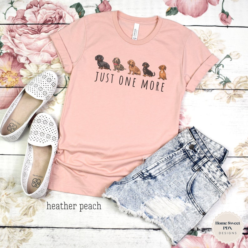Puede incluir: Una camiseta de color melocot&oacute;n con una l&iacute;nea de cinco perros salchicha de diferentes colores y patrones. El texto "Just One More" est&aacute; impreso debajo de los perros.
