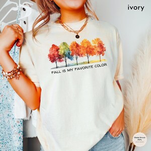 May include: A white t-shirt with a watercolor design of five trees with fall colors. The text "FALL IS MY FAVORITE COLOR" is printed below the trees.