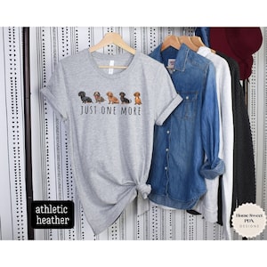Puede incluir: Una camiseta gris con una l&iacute;nea de perros salchicha y el texto "JUST ONE MORE".