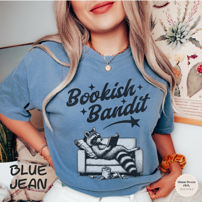 May include: A blue jean t-shirt with the text "Bookish Bandit" in black lettering. A cartoon raccoon wearing a blue shirt is sitting on a couch reading a book. The raccoon has a black and white striped tail.