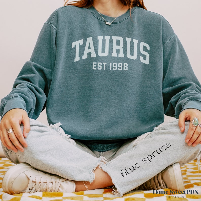 May include: A teal blue sweatshirt with the text "TAURUS EST 1998" in white. The sweatshirt is being worn by a person sitting down with their legs crossed.