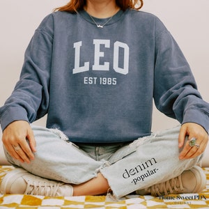 Puede incluir: Una sudadera azul marino con la palabra "LEO" en letras blancas y el texto "EST 1985" debajo.