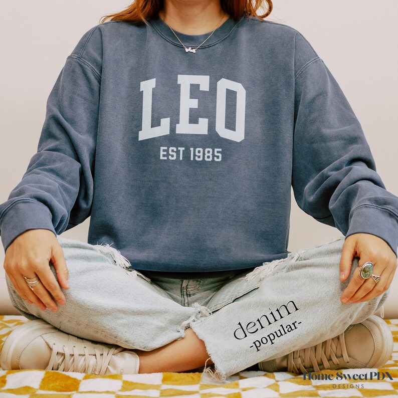 Puede incluir: Una sudadera azul marino con la palabra "LEO" en letras blancas y el texto "EST 1985" debajo.