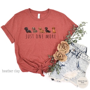 Puede incluir: Una camiseta de color arcilla con una l&iacute;nea de cinco perros salchicha y el texto "JUST ONE MORE".