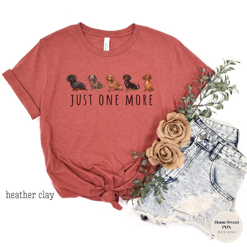 Puede incluir: Una camiseta de color arcilla con una l&iacute;nea de cinco perros salchicha y el texto "JUST ONE MORE".
