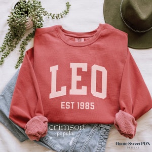 Puede incluir: Sudadera de color rojo carmes&iacute; con letras blancas que dicen "LEO EST 1985".