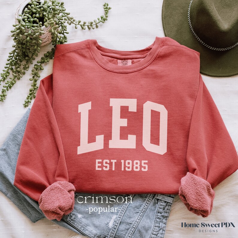 Puede incluir: Sudadera de color rojo carmes&iacute; con letras blancas que dicen "LEO EST 1985".