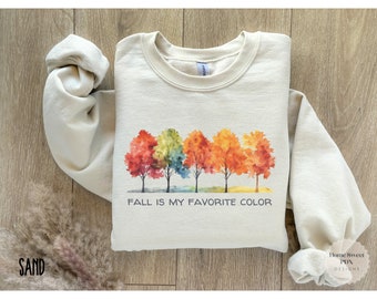 May include: A beige sweatshirt with a watercolor design of five trees with colorful leaves. The text "FALL IS MY FAVORITE COLOR" is printed below the trees.