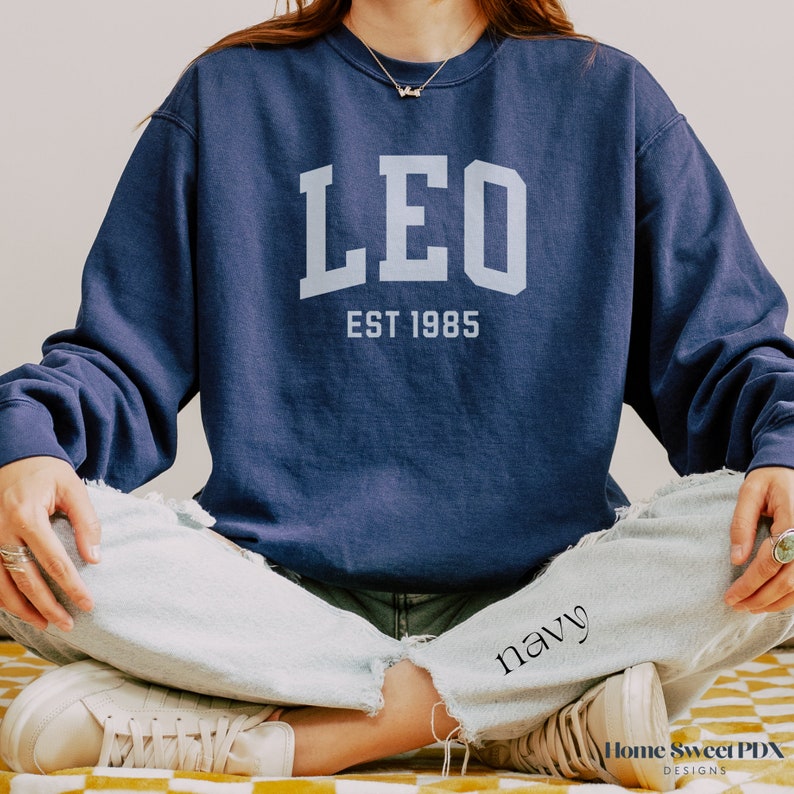 Puede incluir: Una sudadera azul marino con la palabra "LEO" en letras blancas y el texto "EST 1985" debajo.