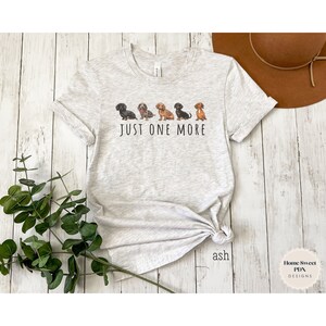 Puede incluir: Una camiseta gris con una l&iacute;nea de cinco perros salchicha de diferentes colores y patrones. El texto "JUST ONE MORE" est&aacute; impreso debajo de los perros.