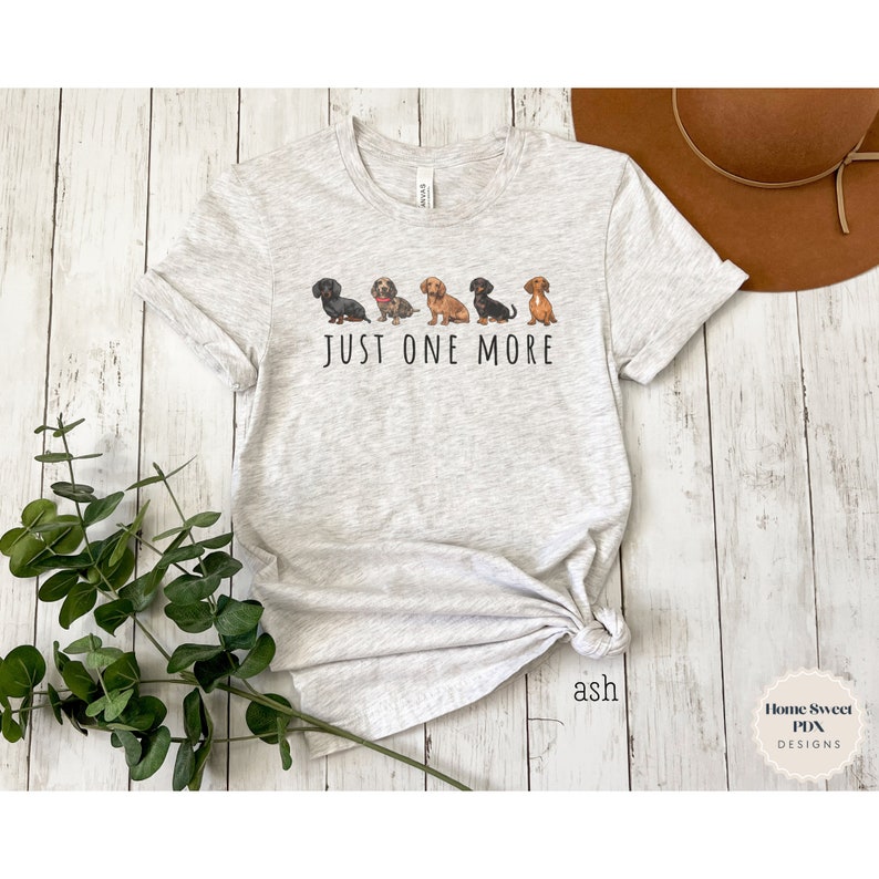 Puede incluir: Una camiseta gris con una l&iacute;nea de cinco perros salchicha de diferentes colores y patrones. El texto "JUST ONE MORE" est&aacute; impreso debajo de los perros.