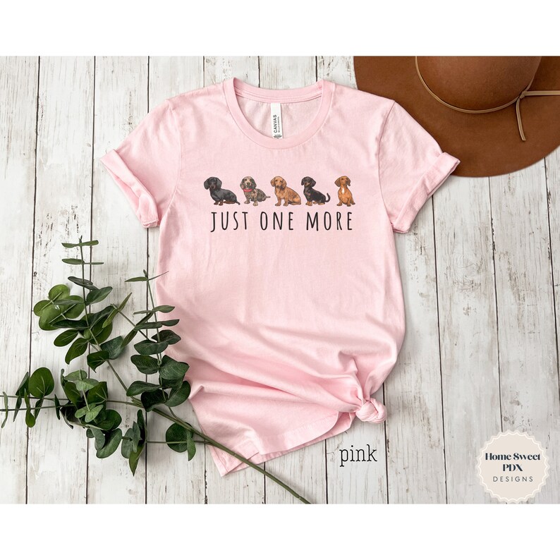 Puede incluir: Camiseta rosa con una l&iacute;nea de perros salchicha y el texto "Just One More".
