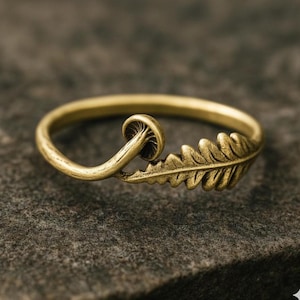 May include: A gold-coloured ring with a mushroom and leaf design. The ring has a smooth band and detailed botanical elements. The mushroom is on the left, and the leaf is on the right. The ring is set against a textured, dark background.