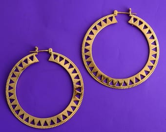 Peut inclure: Deux boucles d'oreilles créoles dorées avec un motif géométrique. Chaque boucle d'oreille présente une série de découpes triangulaires le long du bord extérieur et une petite bordure perlée. Les boucles d'oreilles sont posées sur un fond violet uni.