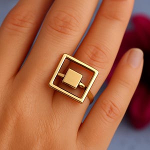 Könnte beinhalten: Ein goldfarbener Ring mit einem geometrischen Design. Der Ring hat ein Quadrat in einem Quadrat, verbunden durch einen horizontalen Balken. Der Ring wird am Finger getragen und zeigt seinen modernen Stil.