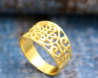 May include: A gold-colored ring with an openwork design. The ring features a band with a continuous pattern of swirling, floral-like shapes. The ring is made of a shiny, yellow metal and is set against a blurred blue background.