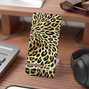 May include: A rectangular phone stand featuring a gold and black leopard print pattern. The stand is made of a durable material and has a slot to hold a phone. The stand is on a wooden desk.