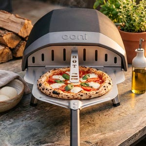 Könnte beinhalten: Ein Pizzaofen mit einer frisch gebackenen Pizza auf einem Metallschieber. Die Pizza hat Tomatenscheiben, Mozzarella und Basilikum. Der Ofen ist schwarz und silberfarben, mit dem Markennamen "ooni" sichtbar. Eine Flasche Olivenöl und eine Schüssel mit Teigbällchen befinden sich in der Nähe.