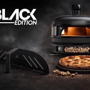 Könnte beinhalten: Schwarzer Pizzaofen der Black Edition mit Pizza und Pizzaschieber. Der Ofen hat eine schwarze Oberfläche, einen Schornstein und einen Holzstauraum. Die Pizza liegt auf einem Holzbrett. Der Pizzaschieber ist schwarz.
