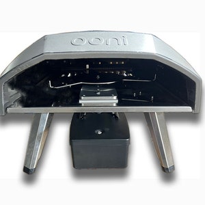 May include: A silver Ooni pizza oven with a black base and stainless steel legs. The oven is open, revealing the interior with a black cooking surface and a metal rack.