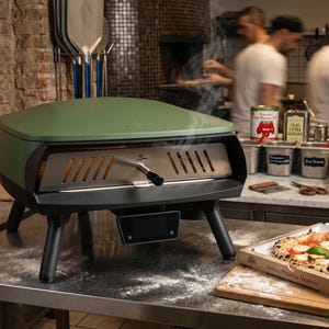 May include: A green and black pizza oven with a stainless steel interior and a handle. A pizza in a box sits on a wooden board. Flour and ingredients are visible in the background.