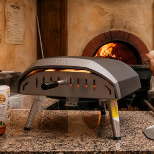 Puede incluir: Un horno para pizza Ooni gris y plateado con un asa negra. Se ven llamas dentro del horno. Una bolsa de harina Caputo y un cuenco de albahaca están en primer plano. Una persona está al fondo.