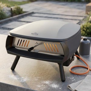 Puede incluir: Un horno de pizza para exteriores gris y negro con interior de acero inoxidable, colocado sobre una superficie de hormigón. El horno tiene un asa, una tubería de gas y está soportado por patas negras. Se ve un pequeño depósito de propano.