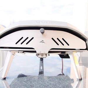 Puede incluir: Un horno de pizza plateado y negro con un diseño de leña. El horno tiene una pequeña puerta en la parte delantera y un logotipo que dice "Ooni".