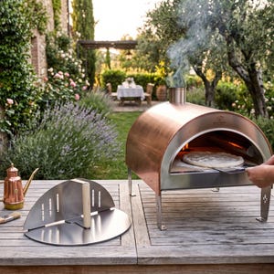 Puede incluir: Un horno de pizza para exteriores de cobre y acero inoxidable con una pizza dentro. Una persona está usando una pala para pizza para colocar la pizza en el horno. Una puerta de horno de pizza de acero inoxidable y un dispensador de aceite de cobre están sobre la mesa de madera.