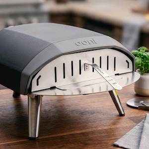 Op de afbeelding: Een zwarte en grijze pizza-oven voor buiten met de merknaam "ooni" op de voorkant. De oven heeft een roestvrijstalen voorkant met verticale ventilatieopeningen en een scharnierende deur. Hij staat op vier zilveren poten en staat op een houten oppervlak.