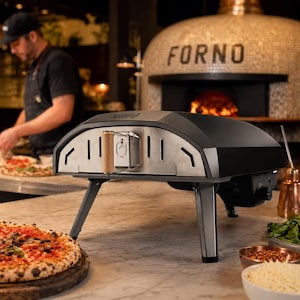 Peut inclure: Un four à pizza extérieur noir et argenté avec une façade en acier inoxydable et un dessus noir. Le four est posé sur un comptoir en marbre, avec une pizza et des ingrédients au premier plan. Une personne prépare une pizza en arrière-plan.