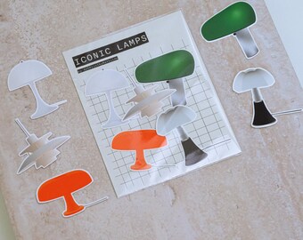 May include: A collection of lamp stickers in various colors and designs, including white, green, and orange. The stickers are arranged on a light-colored surface, with a package of stickers that reads "ICONIC LAMPS".
