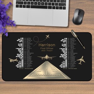 May include: A rectangular black desk mat featuring a gold-colored airplane graphic, a runway illustration, and the text Harrison First Officer 2019-Present. The design includes the NATO phonetic alphabet, Morse code, and small airplane icons.